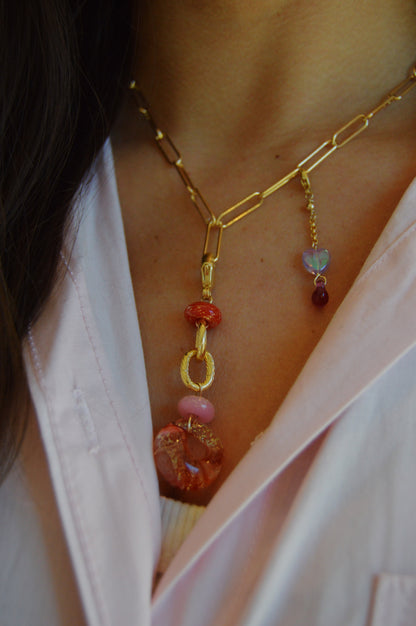 A close-up of a person wearing the Paperclip Layer gold chain necklace with pink, orange, and red beads and gemstone charms, styled over a light pink collared shirt.
