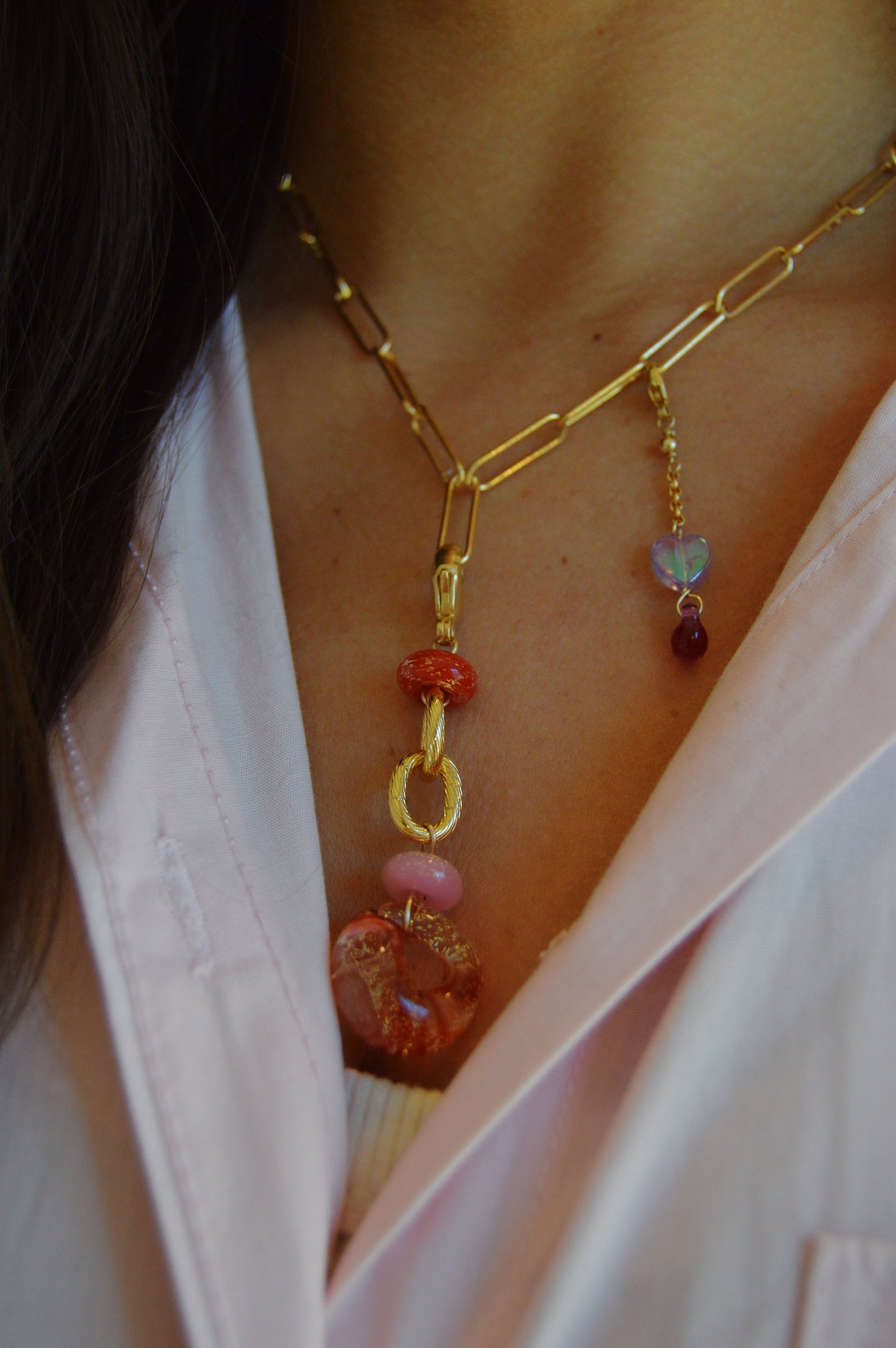 A close-up of a person wearing the Valentine Grand Bubble Charm gold chain necklace with pink and red gemstone pendants, chunky links, and colorful, textured charms, paired with a light pink collared shirt.
