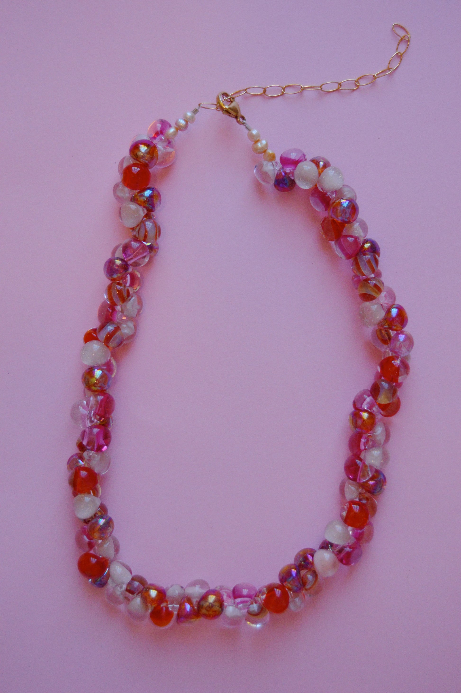 The Valentine Bubble Glass Necklace- Lovebug, with red, pink, purple, and clear handmade beads, gold chain extender, and white pearls, is beautifully displayed on a pastel pink background.