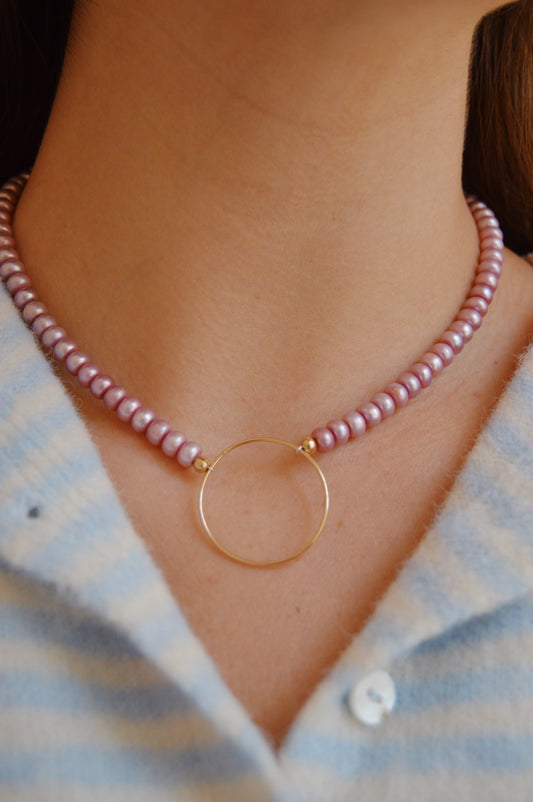 A close-up of a person wearing the Circle Pearl Layer: a handcrafted purple beaded necklace with a 14k gold-filled circular pendant, paired with a light blue and white striped shirt.