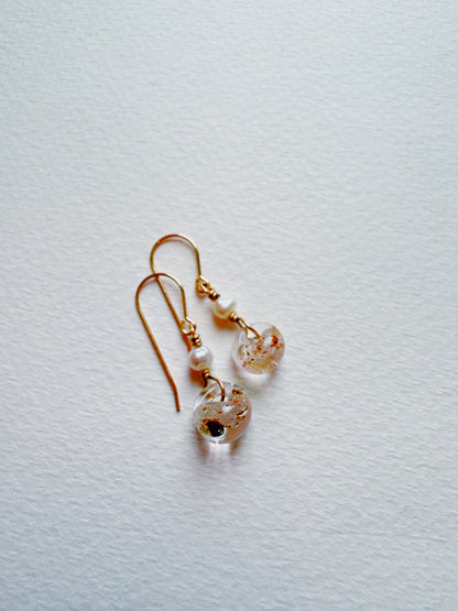 The Bubble Minis are gold hook earrings with small freshwater pearls and round glass beads featuring gold and dark flecks, shown on a textured white surface.