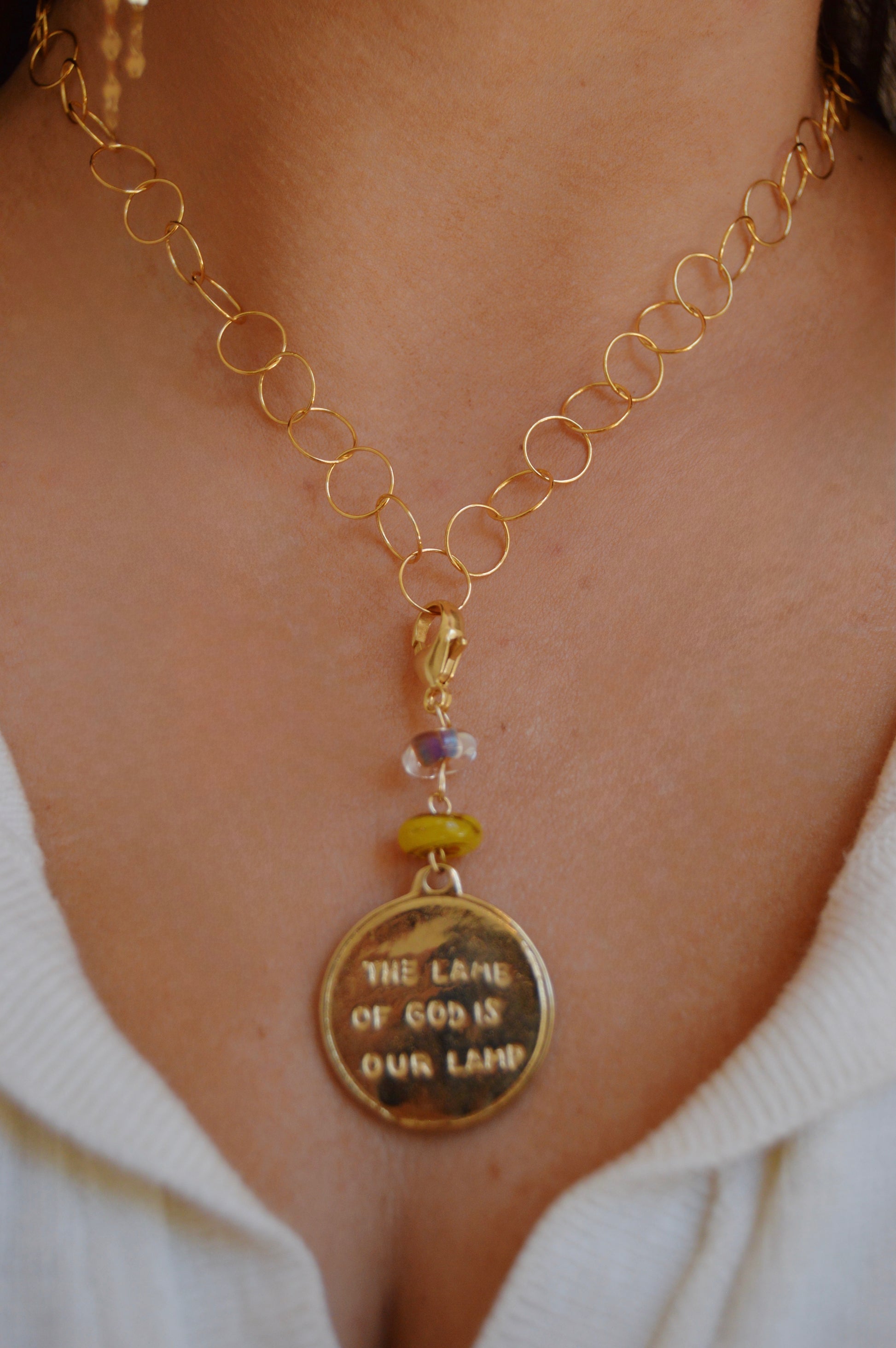 A close-up of the Lamb of God Charm, a gold chain necklace with a round engraved pendant, worn with a cream top. The artisan piece features handmade lampwork glass beads for added elegance.