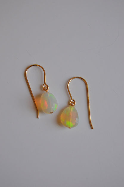 The Baby Opal Drops are 14k gold-filled hook earrings featuring oval, translucent opals with subtle rainbow hues, beautifully displayed on a white background—an elegant showcase of handcrafted jewelry.