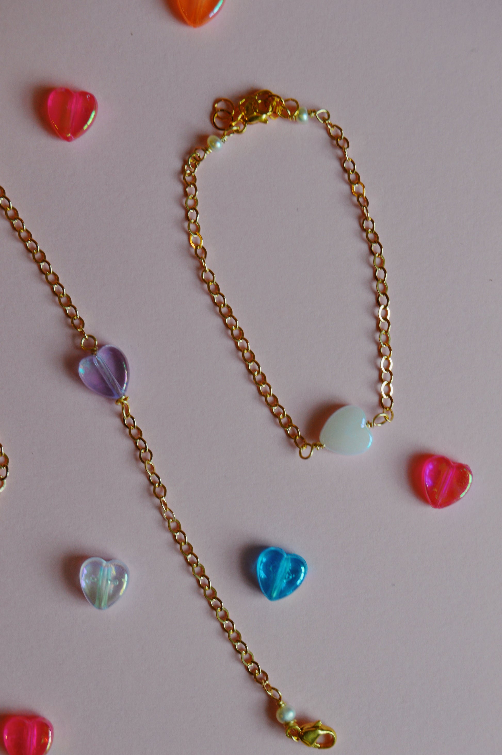 The Friendship Bracelet features gold chain with acrylic heart beads, displayed on a light pink background and surrounded by colorful heart-shaped decorations.