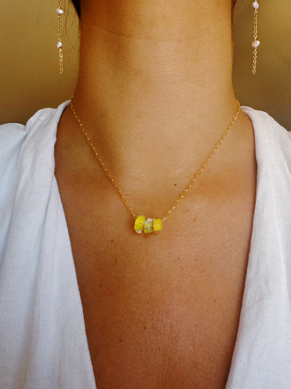 A person wearing the Dainty Donut Layer necklace—three yellow glass donut beads with two silver spacer beads on a 14K gold chain—paired with gold dangling earrings and a white top. Only the neck and upper chest are visible.