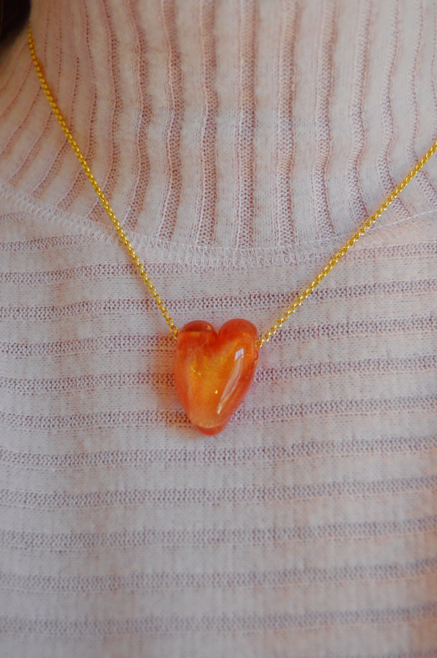 A person wearing the Glass Heart Necklaces- Reds & Oranges, featuring a gold-filled chain and an orange handmade glass heart pendant, shown over a light pink ribbed turtleneck sweater.