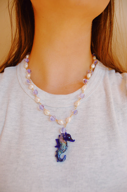 A person wears the Glass Seahorse Layer necklace, featuring cultured freshwater pearls, purple beads, and a blue glass seahorse pendant, paired with a white ribbed shirt. The focus is on the necklace and lower face and neck.