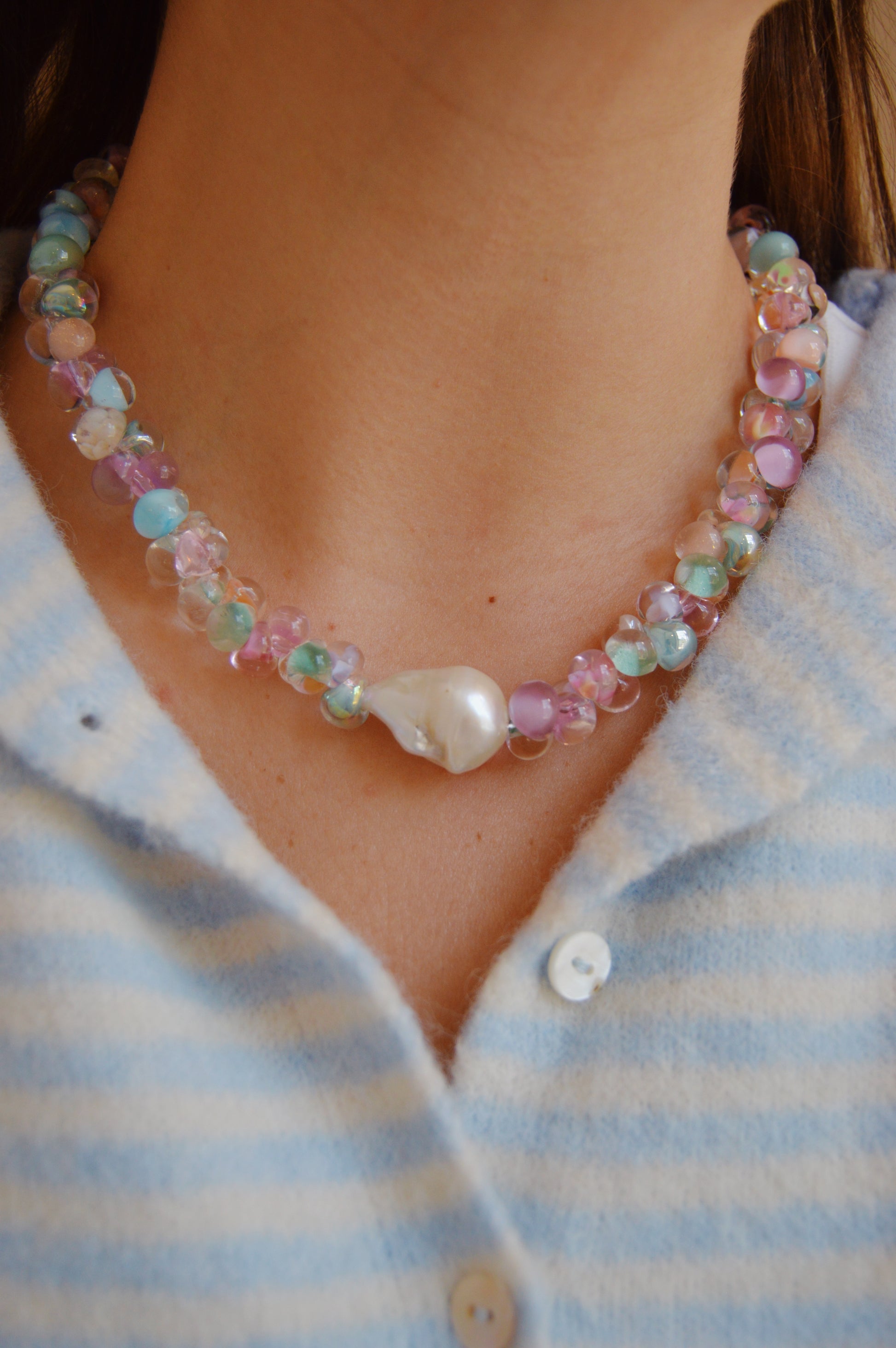 A close-up of someone wearing the Bubble Glass Necklace- Spring Mix, featuring colorful handmade glass beads and a large white pearl pendant, paired with a blue and white striped button-up shirt in soft textured fabric.