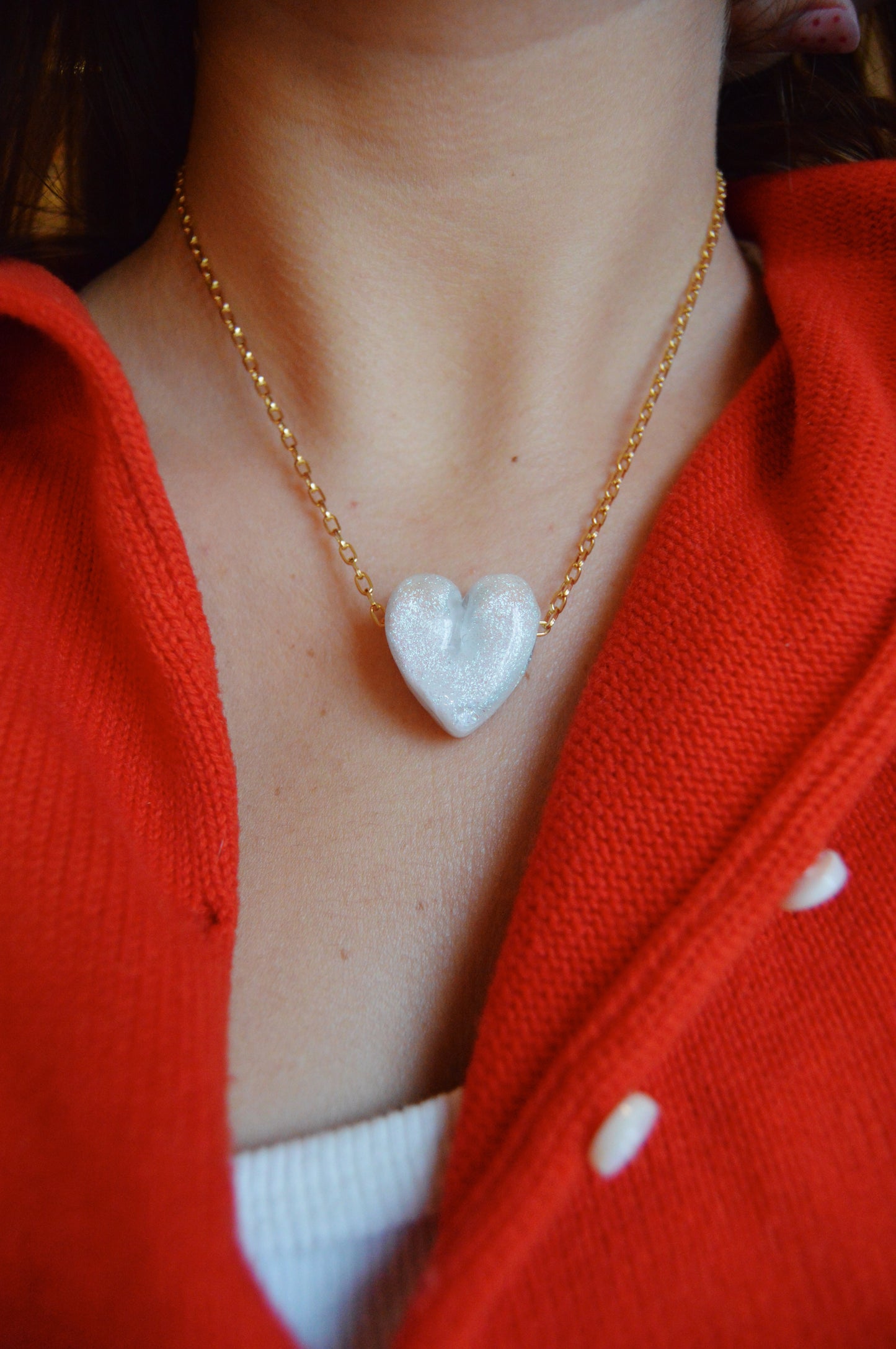 A close-up of a person wearing the Glass Heart Necklaces- Whites and Clears, featuring a handmade lampwork glass heart pendant on a gold-filled chain. The person is dressed in a red top layered over a white shirt.