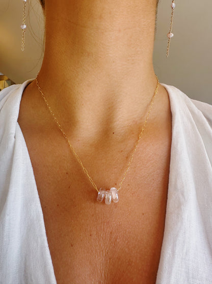 A person wearing the Dainty Donut Layer, a delicate 14K gold chain necklace with translucent pink beads and matching earrings, styled with a white V-neck top. The necklace features an elegant, secure gold-filled clasp.