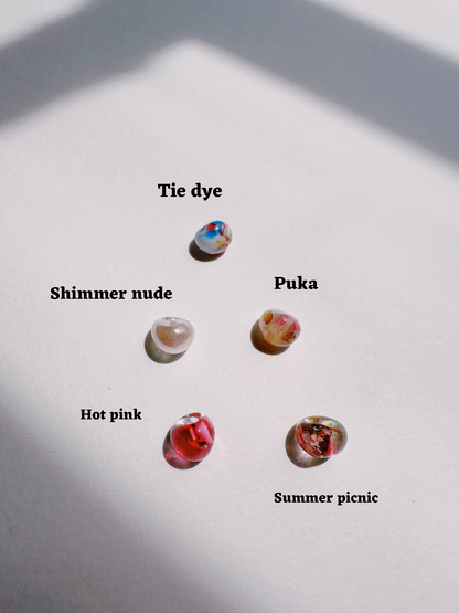 Five small, glossy, oval-shaped handblown glass Bubble Drops beads—Tie dye, Shimmer nude, Puka, Hot pink, and Summer picnic—are displayed on a white surface with shadows.