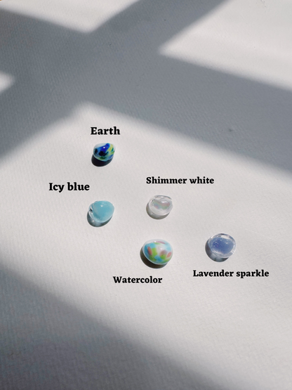 Five handblown Bubble Drops marbles—Earth, Icy Blue, Watercolor, Shimmer White, and Lavender Sparkle—rest on a white surface. Sunlight and shadows emphasize the artistry of these unique glass beads.