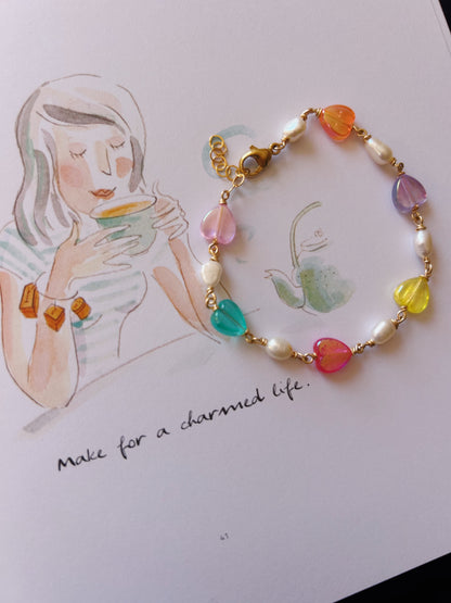 The Bestie Bracelet, featuring colorful acrylic hearts, pearls, and 14K gold filled accents, is displayed on a page illustrated with a woman holding a cup and the phrase "Make for a charmed life.