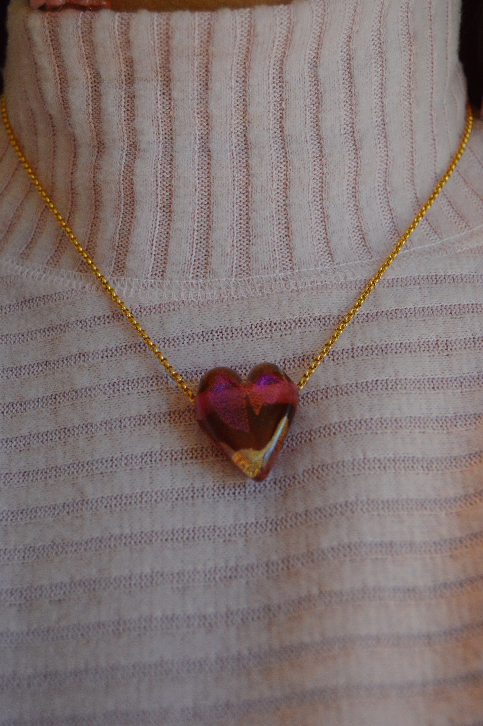 Someone wears a pale pink ribbed turtleneck and an artisan-made gold-filled chain necklace featuring the Glass Heart Necklaces- Reds & Oranges, with a handmade lampwork glass heart-shaped translucent pink pendant.