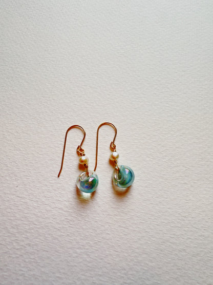Bubble Minis are delicate earrings with gold hooks and gold filled findings, each adorned with a small white freshwater pearl and a round translucent blue-green glass bead, shown on a textured white surface.