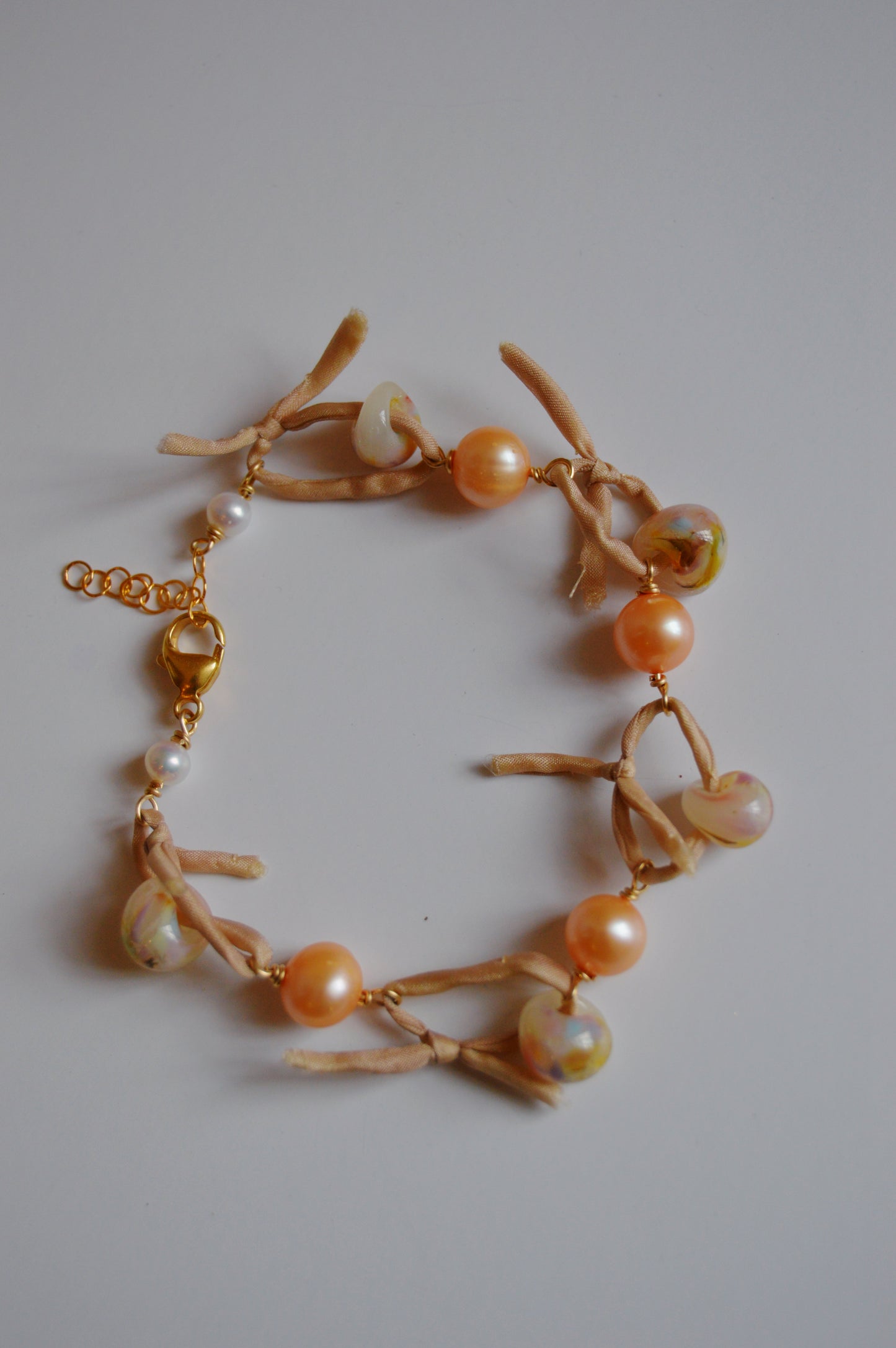 The Silk and Pearl Bracelet- Peachy Puka features peach and marble-patterned beads, light tan suede ties, and a gold clasp, creating an elegant handmade accessory displayed on a plain white background.