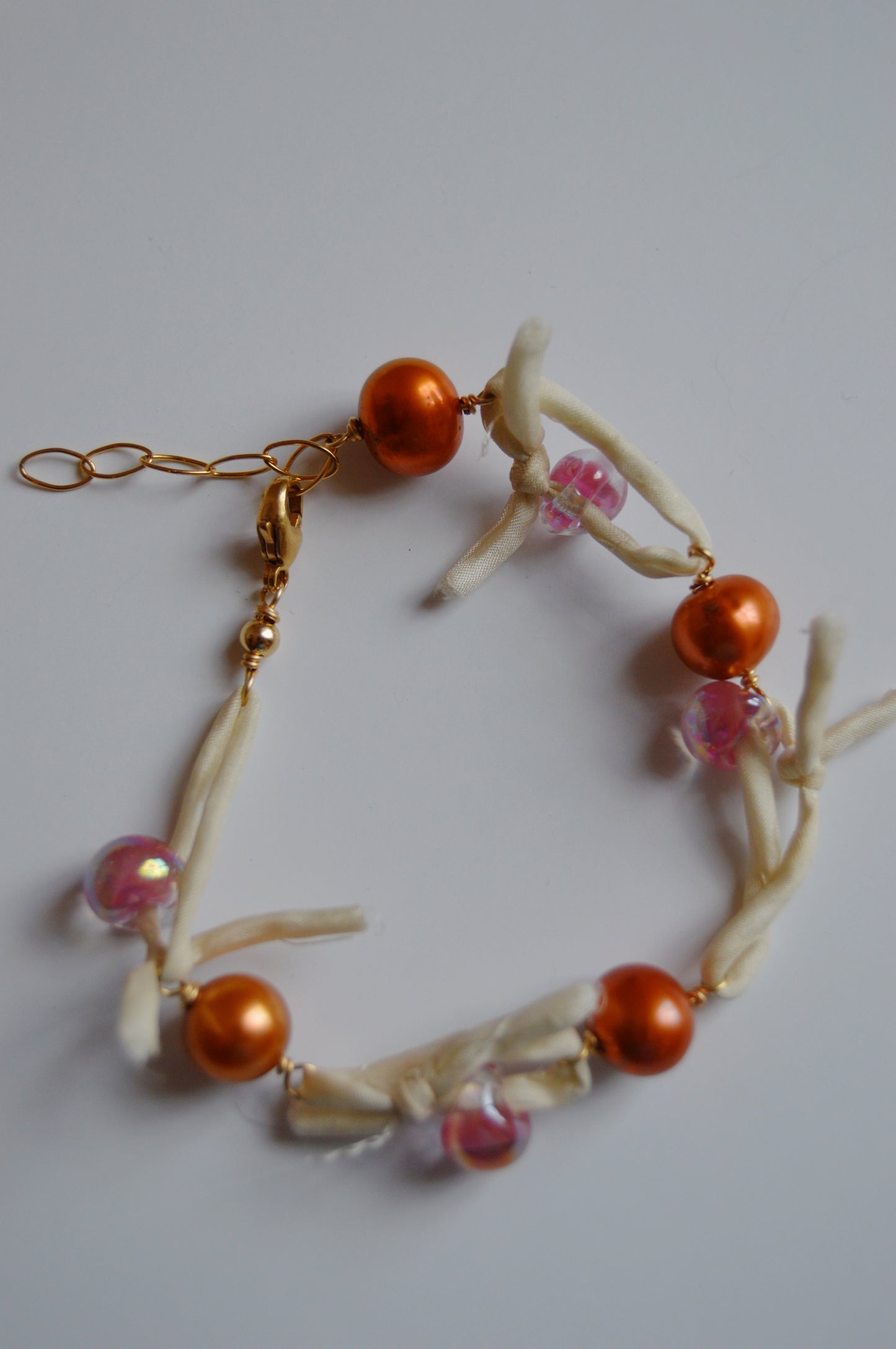 The Silk and Pearl Bracelet- Shimmer Copper showcases orange and pink beads, freshwater pearls, white knotted fabric, and a gold chain clasp, all handmade and displayed on a white background.