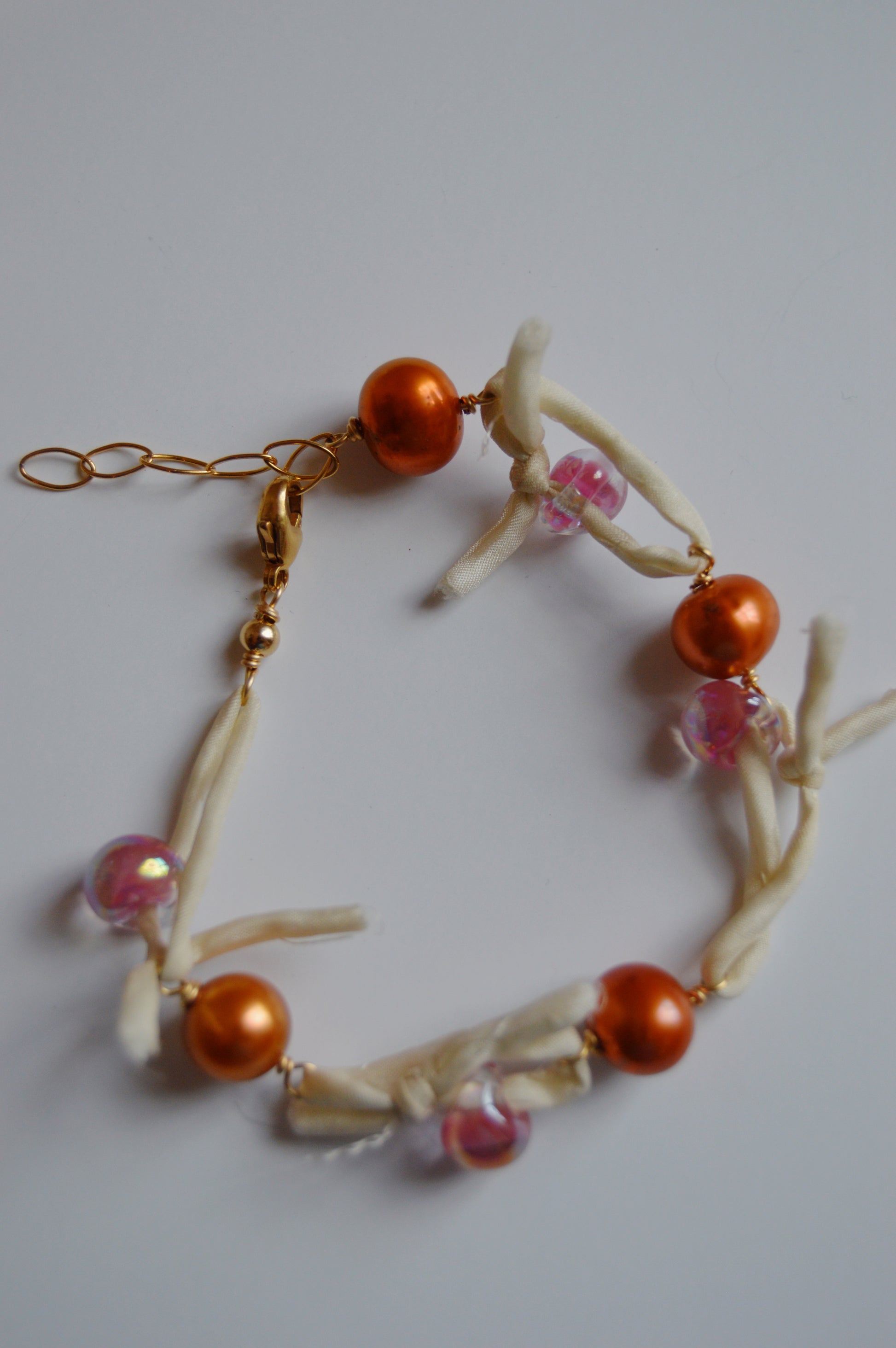 The Silk and Pearl Bracelet- Shimmer Copper showcases orange and pink beads, freshwater pearls, white knotted fabric, and a gold chain clasp, all handmade and displayed on a white background.