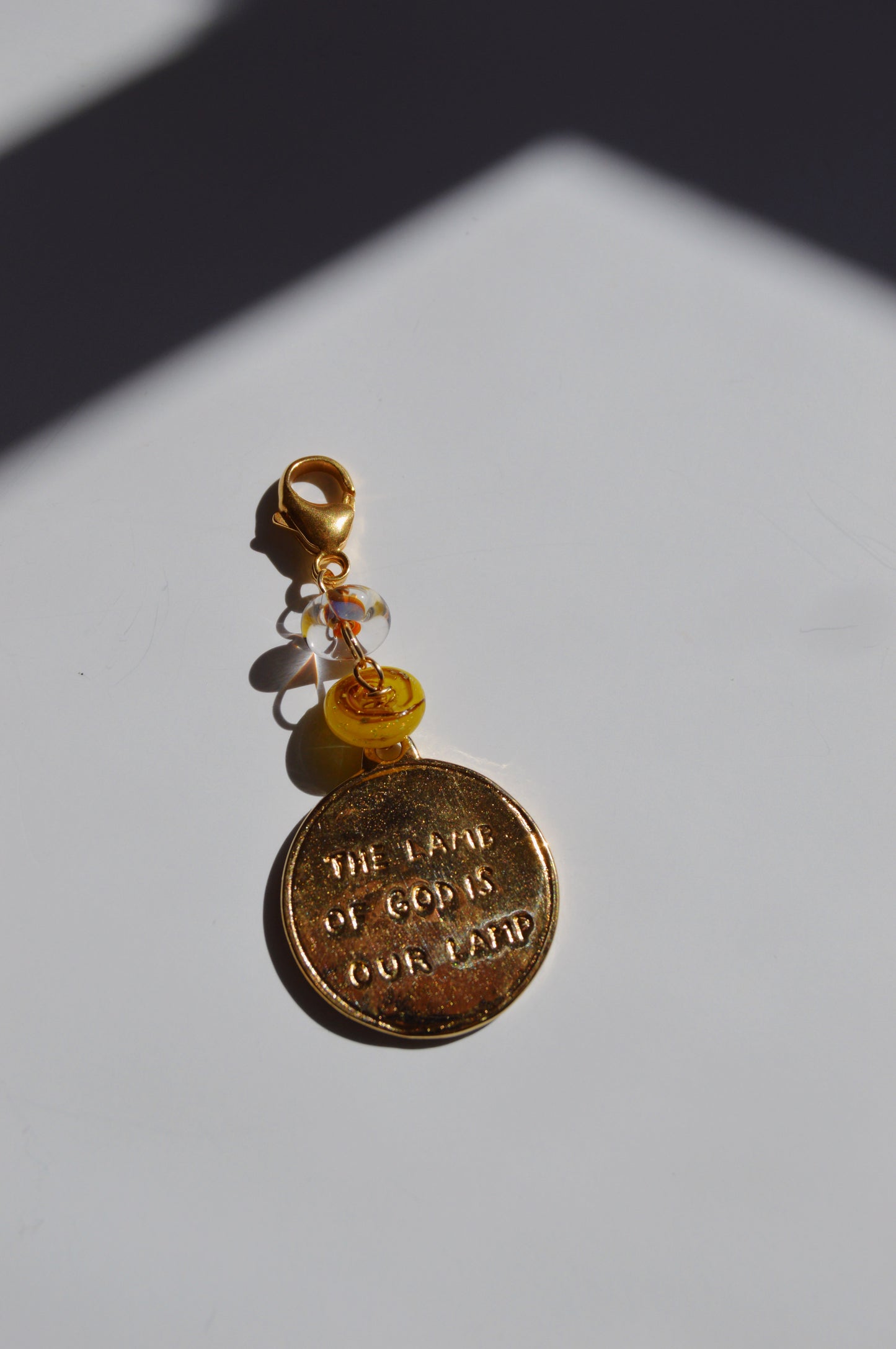 The Lamb of God Charm keychain features a gold medallion engraved with THE LOVE OF GOD IS OUR BAND, 14k gold-filled findings, yellow and clear handmade lampwork glass beads, and a lobster clasp.