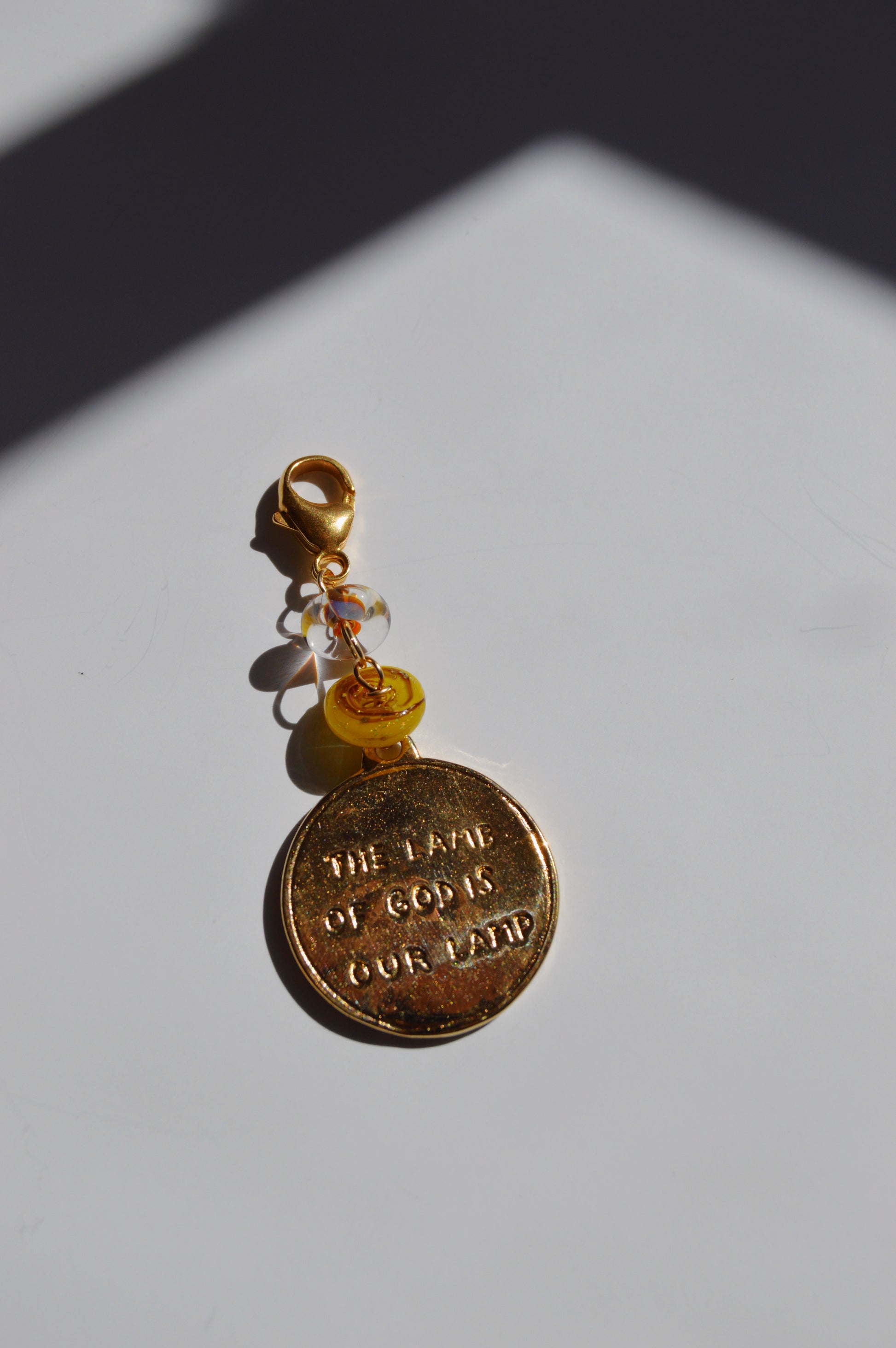 The Lamb of God Charm keychain features a gold medallion engraved with THE LOVE OF GOD IS OUR BAND, 14k gold-filled findings, yellow and clear handmade lampwork glass beads, and a lobster clasp.