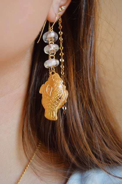 Close-up of a person's ear wearing the Fish and Glass Drops: a 14k gold-filled earring with a Cloisonne fish charm and three handmade lampwork glass beads, shown hanging beside long brown hair.