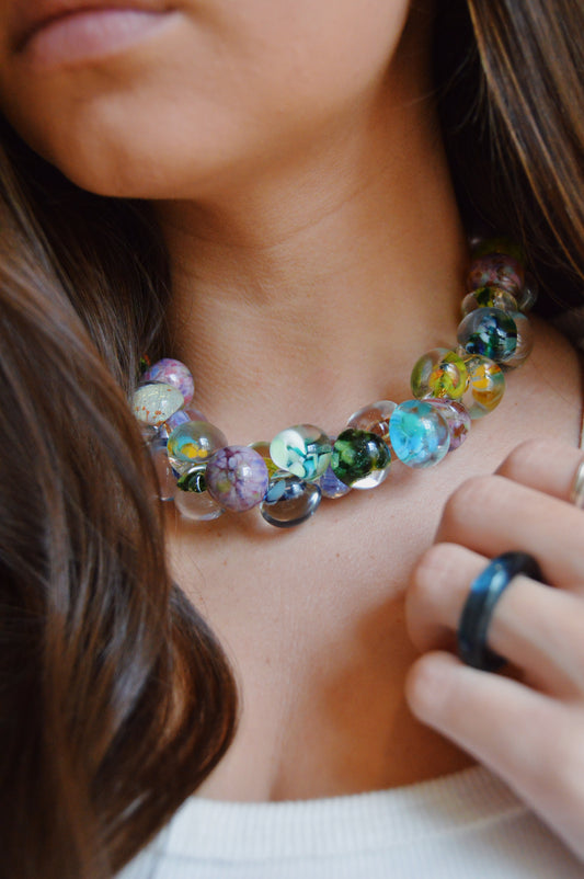A close-up of a woman with long brown hair wearing the Grand Bubble Necklace- Garden Mix, a colorful, chunky piece made with handmade lampwork glass beads. She holds the necklace, displaying a dark ring on her finger and wears a white top.