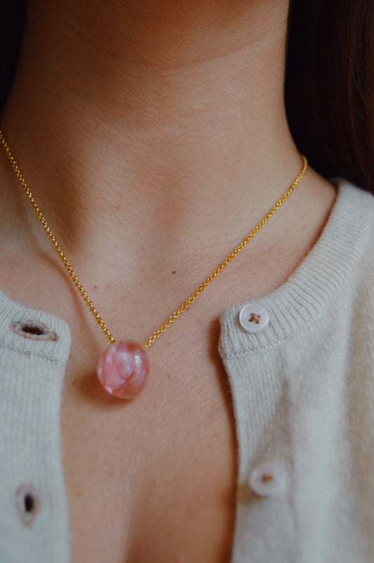 A close-up of a person wearing a beige buttoned cardigan, showcasing the Grand Bubble Single-Petal necklace—an artisan-made piece featuring a 14k gold-filled chain and round pink pendant resting on their collarbone.