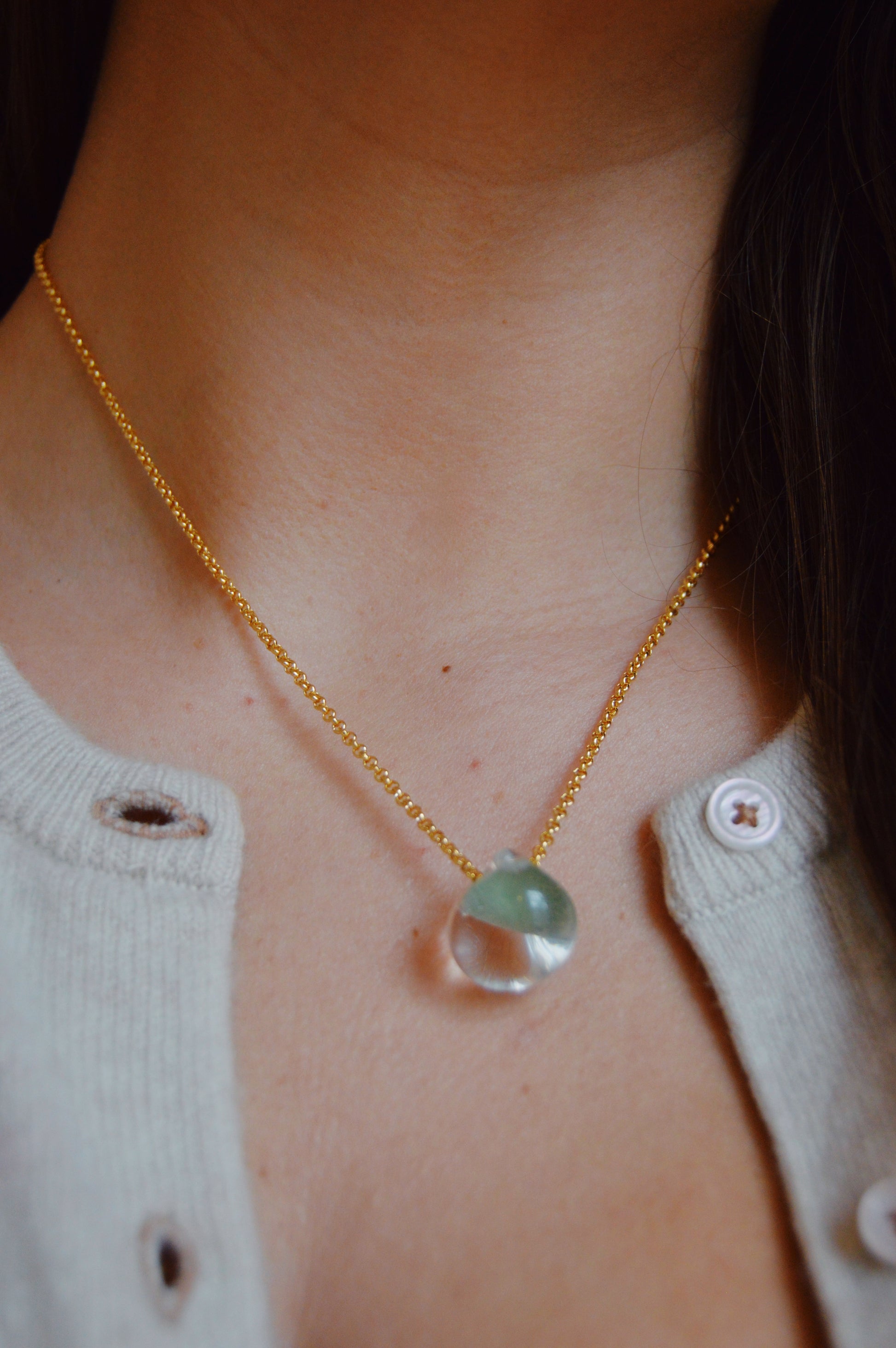 A close-up of a person wearing the Grand Bubble Single- Seafoam, an artisan gold-filled chain necklace featuring a translucent greenish handmade lampwork glass bead pendant, styled with a light beige cardigan and partially visible hair.