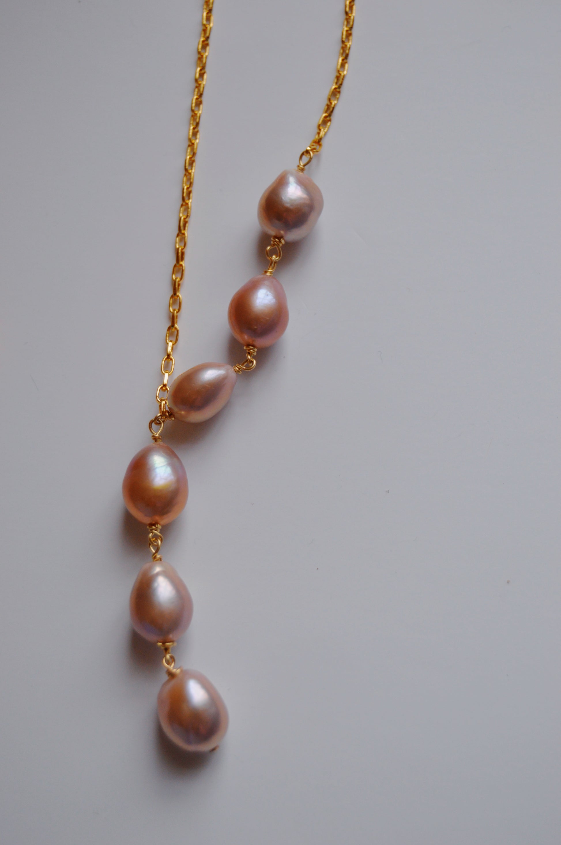 The Champagne Pearl Lariat features six vertically arranged, pink-lavender cultured freshwater pearls on a delicate 14k gold-filled chain—handcrafted for timeless elegance.
