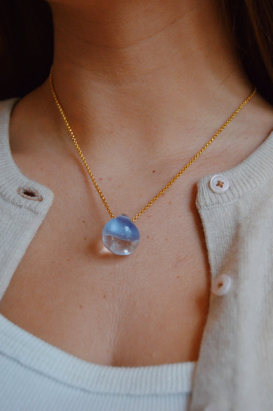 A close-up of a person wearing the Grand Bubble Single- Hydrangea gold chain necklace, featuring a round translucent blue glass bead pendant, styled with a white tank top and light beige cardigan.