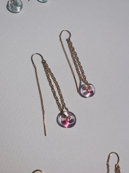 Glass Donut Threaders are delicate 14k gold filled chain earrings with circular, handblown glass beads accented with a touch of pink, shown on a white textured surface.