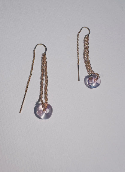 The Glass Donut Threaders are gold chain earrings with handblown glass beads, featuring two 14k gold-filled threader strands and a bar detail at the end, displayed on a textured white surface.