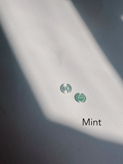 Two small, translucent mint-green Glass Donut Threaders rest on a white surface with soft shadows. The word "Mint" is written nearby, highlighting their gentle color and clarity reminiscent of delicate handblown glass beads.