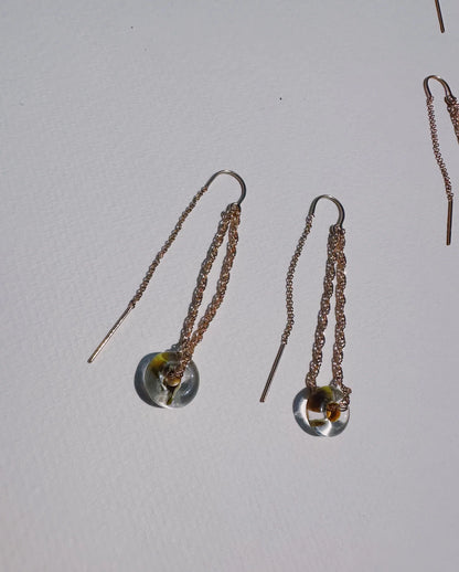 The Glass Donut Threaders are delicate 14k gold filled earrings featuring small circular handblown glass beads with yellow and brown accents and metal rods, displayed on a textured white surface.