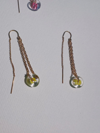 The Glass Donut Threaders, gold chain earrings with translucent circular pendants and small yellow accents, featuring delicate handblown glass beads, are displayed on a white textured surface.