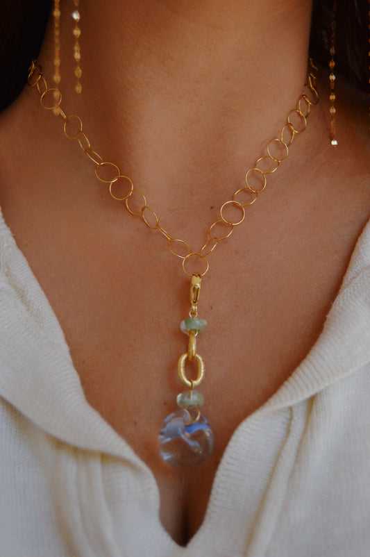 A close-up of a person wearing the Grand Bubble Charm- Periwinkle, a handmade gold chain necklace with circular links, featuring periwinkle lampwork glass beads and a clear stone pendant. Paired with an off-white top for an elegant look.