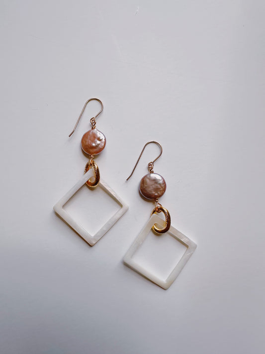 The Diamond Shell and Pearl Drops are dangle earrings with cultured freshwater pearls atop gold hooks, featuring large white square frames suspended below by a small gold loop. Displayed against a plain white background.