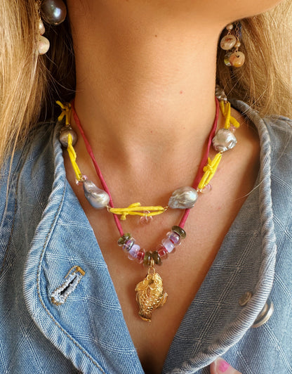 A close-up of a person wearing the Cloisonne Fish Layer - Berry necklace with yellow and pink cords, pearls, handblown glass beads, a gold pendant necklace, a blue textured jacket, and a pearl earring.
