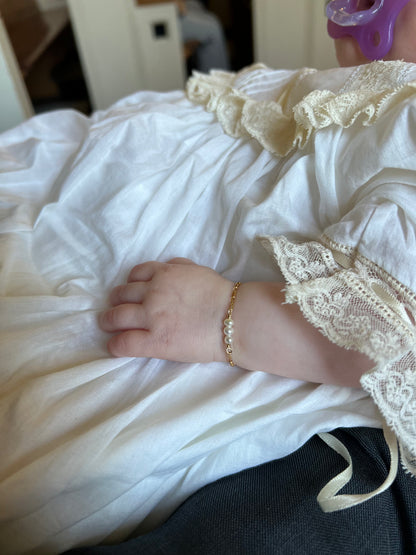 A baby wearing a white lace dress and a Baby Bracelet with cultured freshwater pearls rests their hand on their lap, holding a purple pacifier.