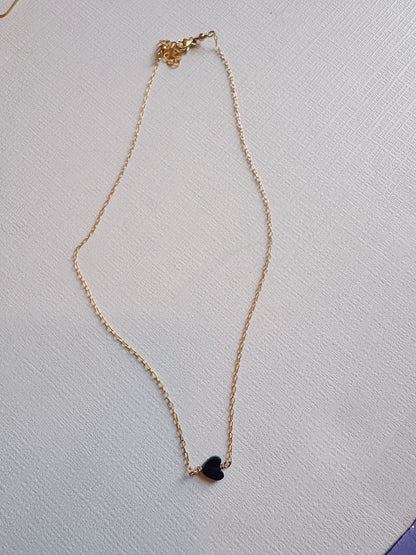 The Boyfriend Necklace, a delicate gold-filled chain with a small black heart pendant, is displayed on a textured white surface.
