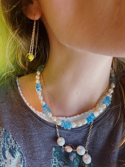 A close-up of a person's neck and lower face wearing Glass Donut Threaders—14k gold filled threader earrings with a small yellow glass bead—alongside blue and white beaded necklaces, against a green background.