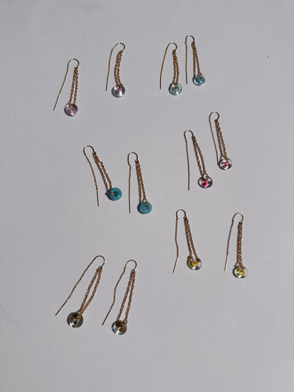 Thirteen Glass Donut Threaders, each featuring small, colorful handblown glass beads in blue, purple, pink, yellow, and clear hues, are arranged on a white surface. One threader is missing its pair.