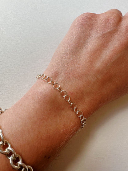 A close-up of a wrist wearing the Dainty Silver Bracelet, a delicate sterling silver chain with round links, alongside a chunkier silver bracelet. The background is plain and light.