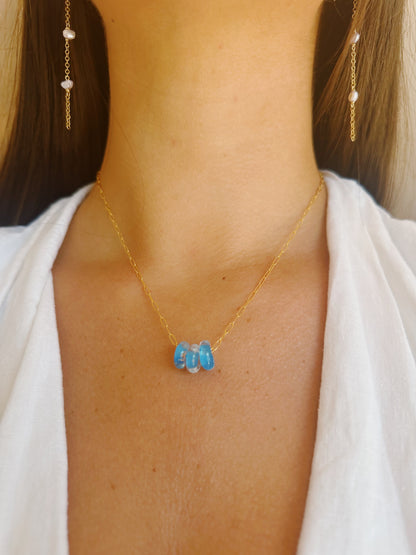 A woman in a white V-neck top wears the Dainty Donut Layer: a 14K gold chain necklace with three blue glass donut beads and matching gold chain earrings with small white beads.