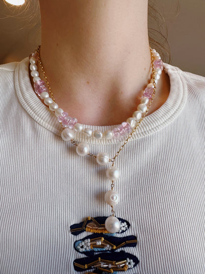 A person wearing the 6 Pearl Lariat - White Pearls necklace layered with pink bead necklaces on a white ribbed shirt featuring a blue and gold embroidered patch at the neckline.