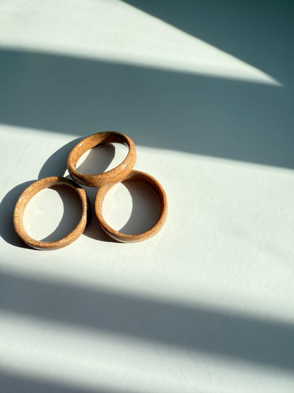 Three Wooden Ring- Light Wood pieces are arranged in a triangle on a white surface, with diagonal light and shadow patterns crossing the background.
