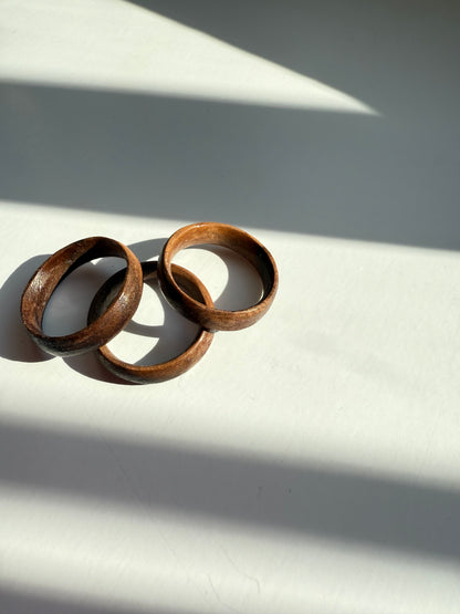 The Wooden Ring- Dark Wood, an artisan-made handmade piece, rests on a white surface with sunlight and shadows casting diagonal patterns across the image.