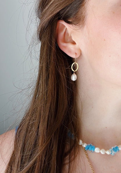 A close-up of a woman's ear wearing Gold Filled Pearl Drops—gold hoop earrings with dangling cultured freshwater pearls. She also wears a 14k gold filled chain necklace with blue and white beads, her brown hair partially covering her ear.