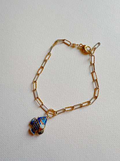 The Fish Anklet is a gold-plated chain with rectangular links, adorned with a blue cloisonné fish charm featuring gold and red details, shown on a lightly textured surface.