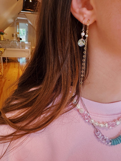 Close-up of a woman in a light pink top wearing Bubble Minis gold chain earrings with freshwater pearls and beaded necklaces featuring glass beads, in a bright, pastel-decorated room with wooden floors.